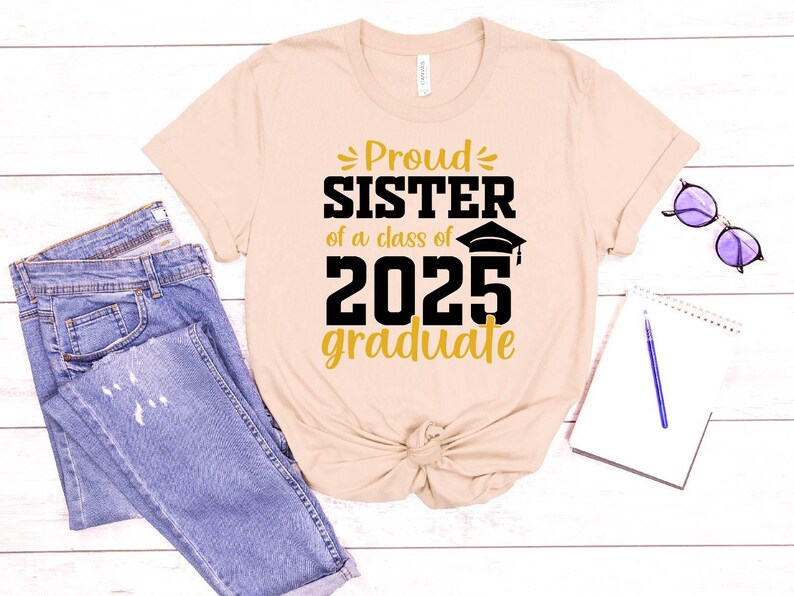 May include: A beige t-shirt with the text "Proud Sister of a class of 2025 graduate" in gold and black lettering. The shirt is laid out on a wooden surface with a pair of jeans and a notebook.