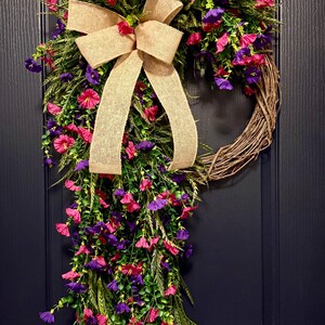 May include: A decorative wreath with a natural grapevine base, adorned with vibrant pink and purple flowers, green foliage, and a large burlap bow. The wreath is displayed on a dark blue door.