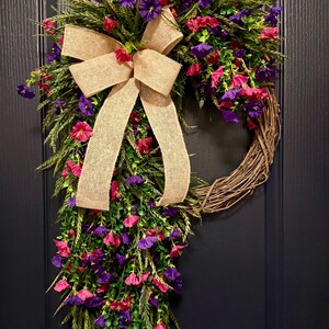May include: A decorative wreath featuring a natural grapevine base, adorned with vibrant pink and purple flowers, green foliage, and a large burlap bow. The wreath is designed for door or wall decor.