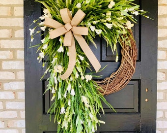 May include: A spring-themed wreath with white flowers and green foliage, accented with a hessian bow. The wreath is hanging on a black door.