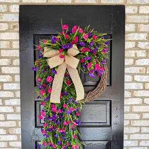 May include: A decorative wreath with a brown grapevine base, adorned with vibrant pink and purple flowers, green foliage, and a tan burlap bow. The wreath hangs on a black door against a brick wall.