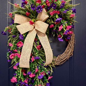 May include: A decorative wreath featuring a natural grapevine base, adorned with vibrant pink and purple flowers, green foliage, and a large burlap bow. This floral wreath is perfect for adding a touch of color to any door or wall.