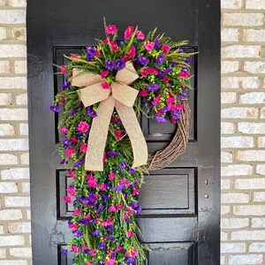 May include: A vibrant floral wreath with pink and purple flowers, green foliage, and a tan burlap bow. The wreath is displayed on a black door against a brick wall, creating a welcoming aesthetic.