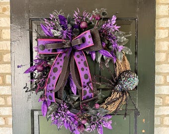 May include: A Halloween wreath with a grapevine base, adorned with purple and black accents. It features a large bow with polka dots, decorative leaves, berries, and a glittery spider. The wreath is displayed on a black door.