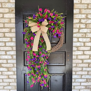 May include: A vibrant, handcrafted wreath with a natural grapevine base, adorned with pink and purple flowers, green foliage, and a beige burlap bow. The wreath is displayed on a black door against a brick wall, creating a welcoming aesthetic.