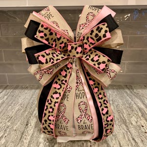 May include: A decorative bow featuring multiple layers of ribbon in various colors and patterns. The bow includes ribbons with leopard print, pink, black, and tan colors. Some ribbons display the words "HOPE" and "be BRAVE" with pink awareness ribbons.