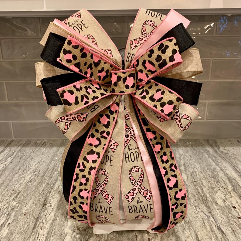 May include: A decorative bow featuring multiple layers of ribbon in various colors and patterns. The bow includes ribbons with leopard print, pink, black, and tan colors. Some ribbons display the words "HOPE" and "be BRAVE" with pink awareness ribbons.