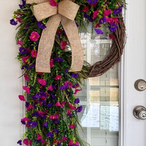 May include: A vibrant floral wreath with a natural grapevine base, adorned with lush greenery and cascading pink and purple flowers. A large burlap bow adds a rustic touch. The wreath hangs on a white door.