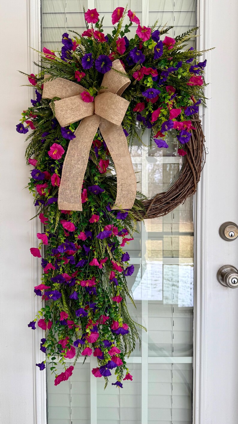 May include: A vibrant floral wreath with a natural grapevine base, adorned with lush greenery and cascading pink and purple flowers. A large burlap bow adds a rustic touch. The wreath hangs on a white door.