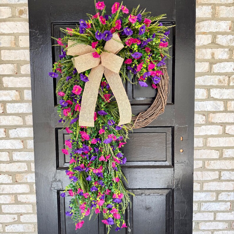 May include: A vibrant floral wreath with pink and purple flowers, green foliage, and a tan burlap bow. The wreath is made of woven twigs and hangs on a black door, creating a welcoming entrance.