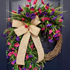 May include: A decorative wreath featuring a natural grapevine base, adorned with vibrant pink and purple flowers, green foliage, and a large burlap bow. The wreath is designed for door or wall decor.