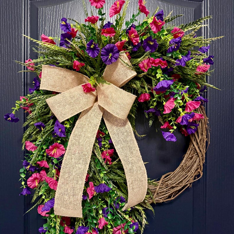 May include: A decorative wreath featuring a natural grapevine base, adorned with vibrant pink and purple flowers, green foliage, and a large burlap bow. The wreath is designed for door or wall decor.
