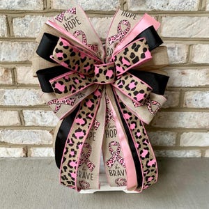 May include: A decorative bow with ribbons in pink, black, and tan. The ribbons feature a leopard print design and the words "Hope" and "Be Brave". A pink ribbon with a breast cancer awareness ribbon is also present.