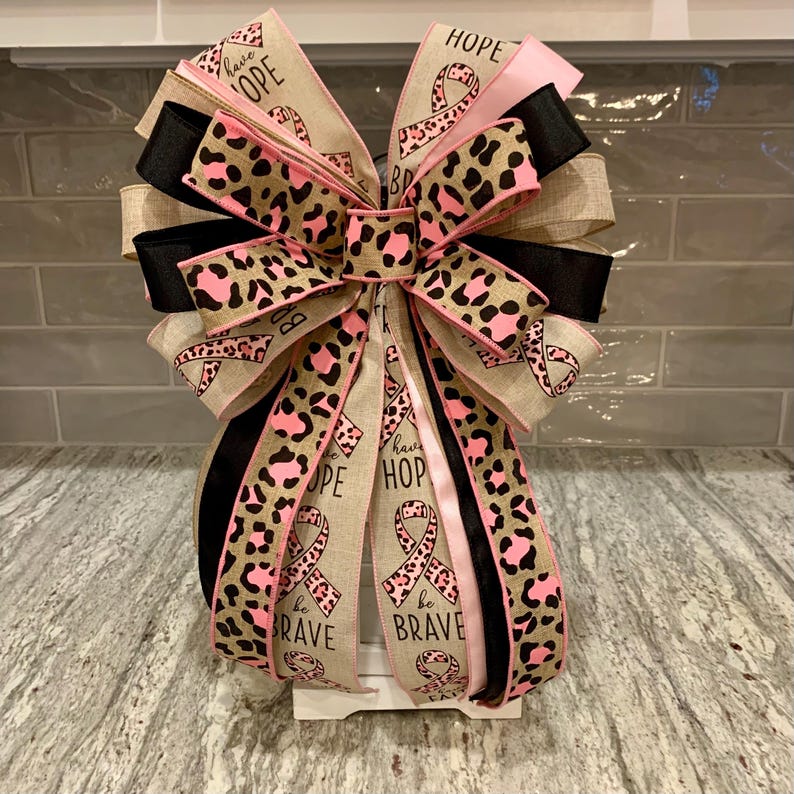 May include: A decorative bow with multiple layers of ribbon in various patterns and colors. The bow features ribbons with leopard print, pink ribbons with breast cancer awareness ribbons, and black and tan ribbons. The words "HOPE" and "BE BRAVE" are visible.