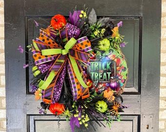 May include: A Halloween wreath featuring a central sign with the text TRICK OR TREAT, surrounded by orange, purple, and green pumpkins, black foliage, and a large multi-patterned bow with orange, purple, and lime green striped and polka dot ribbons.
