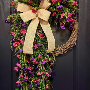 May include: A decorative wreath featuring a grapevine base adorned with vibrant pink and purple flowers, green foliage, and a large burlap bow. The wreath is designed for door or wall decor.