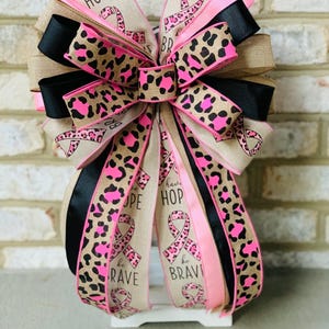 May include: A decorative bow with multiple layers of ribbon. The bow features ribbons with a leopard print design in pink and brown, solid black, and pink ribbons. The bow has the words "HOPE", "be BRAVE", and a pink ribbon symbol.