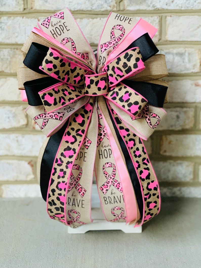 May include: A decorative bow with multiple layers of ribbon. The bow features ribbons with a leopard print design in pink and brown, solid black, and pink ribbons. The bow has the words "HOPE", "be BRAVE", and a pink ribbon symbol.