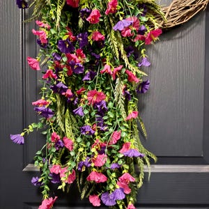 May include: A decorative wreath featuring a woven grapevine base adorned with cascading faux flowers. The flowers are a mix of vibrant pink and purple hues, complemented by green foliage. The wreath is designed for door or wall decor.