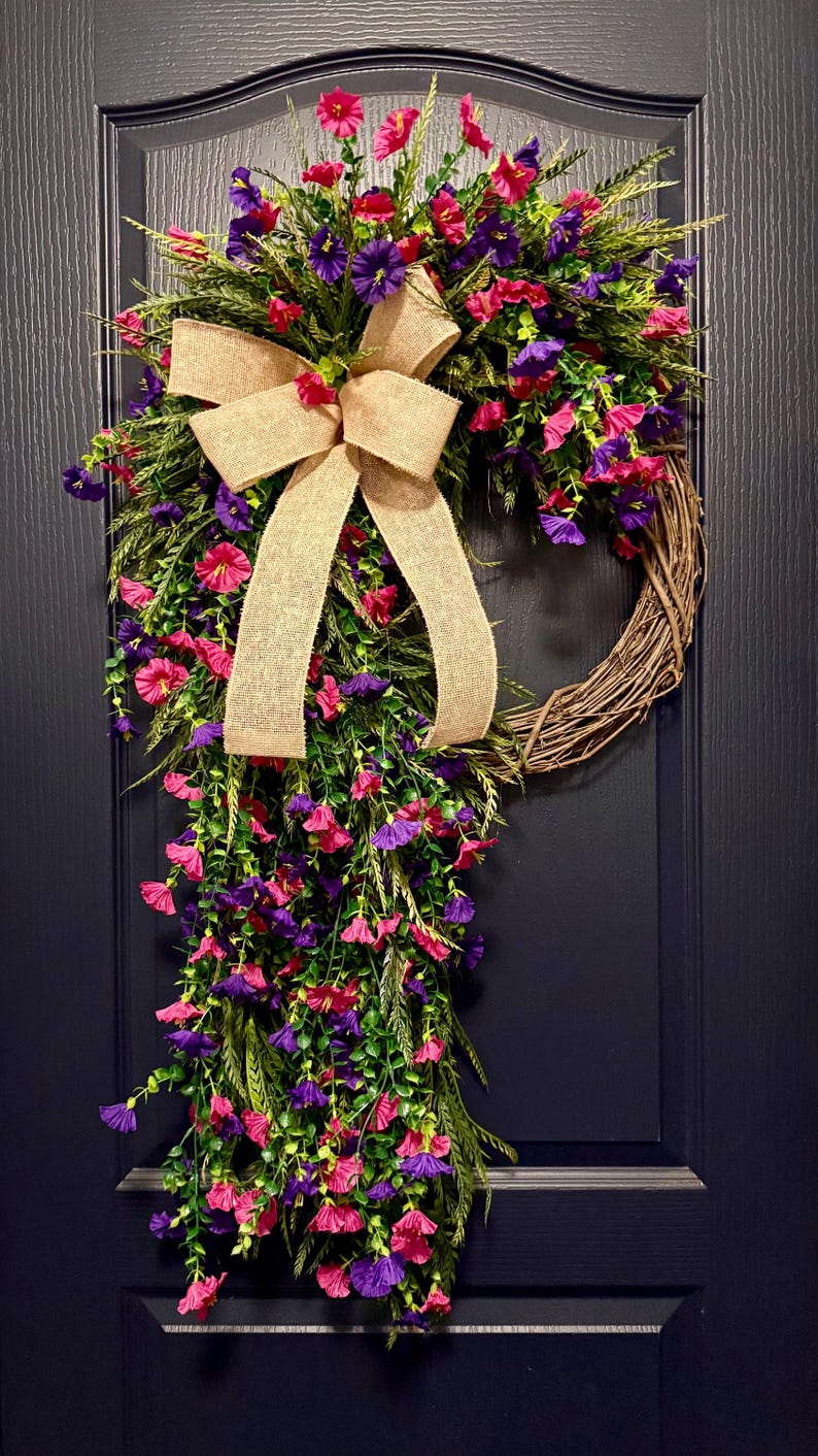 May include: A decorative wreath with a natural grapevine base, adorned with vibrant pink and purple flowers, green foliage, and a large burlap bow. The wreath is displayed on a dark blue door.