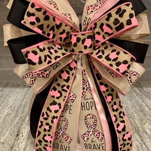May include: A decorative bow featuring multiple layers of ribbon in various patterns and colors. The bow includes ribbons with a leopard print design in pink and brown, solid black and pink ribbons, and ribbons with the words "HOPE" and "be BRAVE" with a pink ribbon symbol.