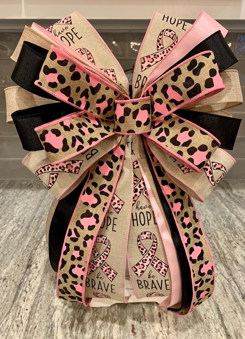 May include: A decorative bow featuring multiple layers of ribbon in various patterns and colors. The bow includes ribbons with a leopard print design in pink and brown, solid black and pink ribbons, and ribbons with the words "HOPE" and "be BRAVE" with a pink ribbon symbol.