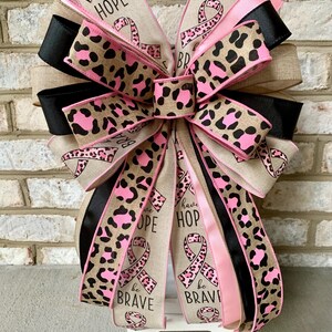 May include: A decorative bow featuring ribbons in various patterns and colors. The bow includes ribbons with leopard print, solid black, and pink. Some ribbons have the words "have HOPE" and "be BRAVE" printed on them, along with a pink ribbon symbol.