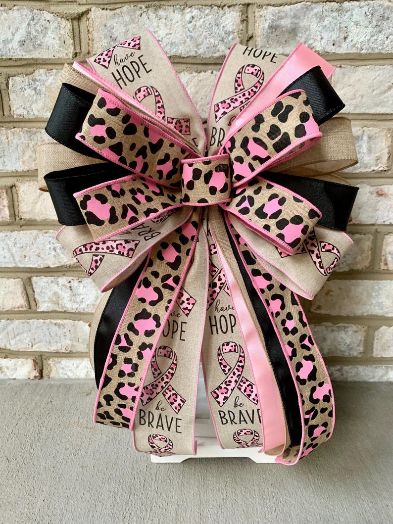 May include: A decorative bow featuring ribbons in various patterns and colors. The bow includes ribbons with leopard print, solid black, and pink. Some ribbons have the words "have HOPE" and "be BRAVE" printed on them, along with a pink ribbon symbol.
