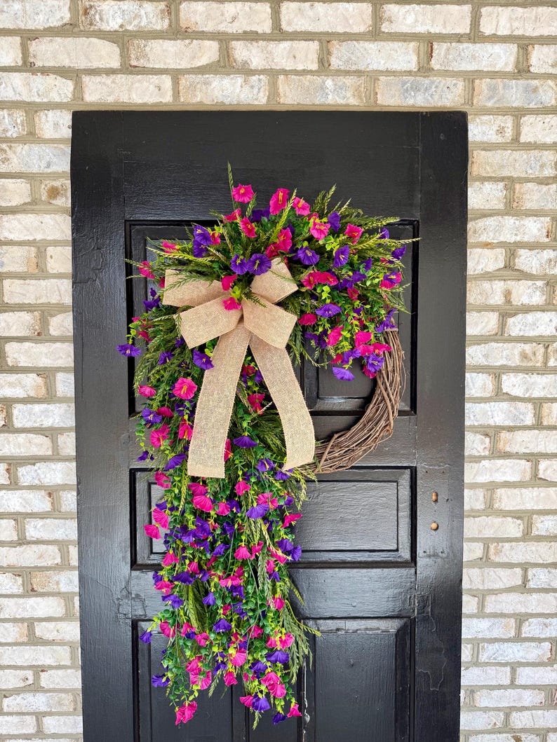 May include: A decorative wreath with a brown grapevine base, adorned with vibrant pink and purple flowers, green foliage, and a tan burlap bow. The wreath hangs on a black door against a brick wall.
