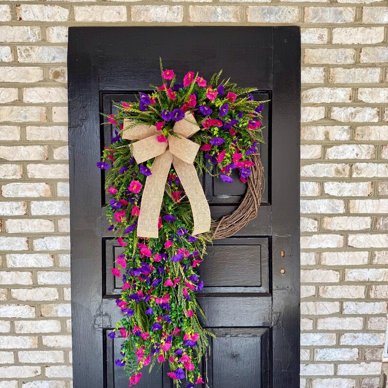 May include: A vibrant floral wreath with pink and purple flowers, green foliage, and a tan burlap bow. The wreath is displayed on a black door against a brick wall, creating a welcoming aesthetic.