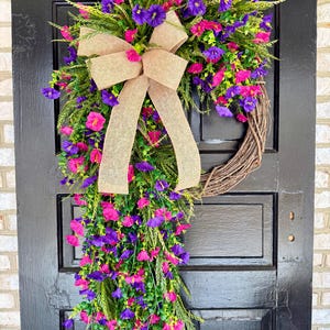 May include: A vibrant floral wreath with a brown grapevine base, adorned with pink and purple flowers, green foliage, and a tan burlap bow. The wreath is displayed on a black door, creating a striking contrast.