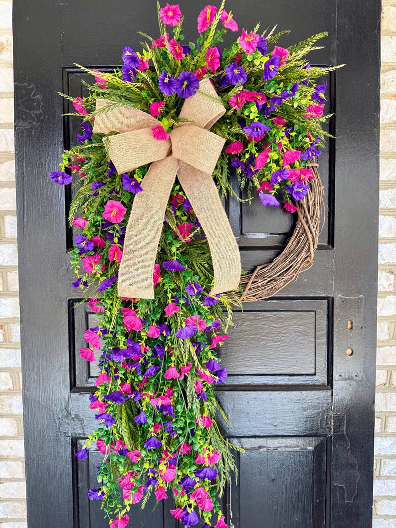 May include: A vibrant floral wreath with a brown grapevine base, adorned with pink and purple flowers, green foliage, and a tan burlap bow. The wreath is displayed on a black door, creating a striking contrast.