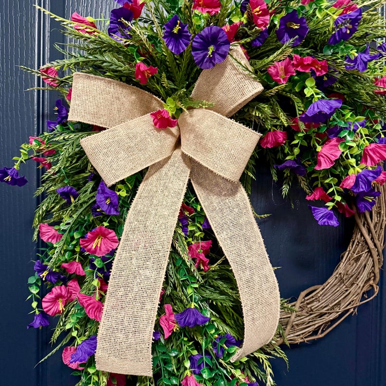 May include: A grapevine wreath adorned with vibrant pink and purple flowers, green foliage, and a large burlap bow. The wreath is a decorative piece, perfect for adding a touch of nature to any home decor. The wreath is approximately 18 inches in diameter.