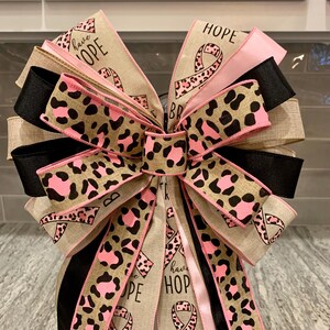 May include: A decorative bow featuring multiple layers of ribbon in various colors and patterns. The bow includes ribbons with a leopard print design in pink and brown, solid black, and tan ribbons. Some ribbons have the words "HOPE" and "have HOPE" printed on them, along with a pink ribbon symbol.