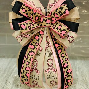 May include: A decorative bow featuring ribbons in pink, black, and tan. The bow has a leopard print pattern in pink and brown. Some ribbons have the words "have HOPE" and "be BRAVE" with a pink ribbon symbol.