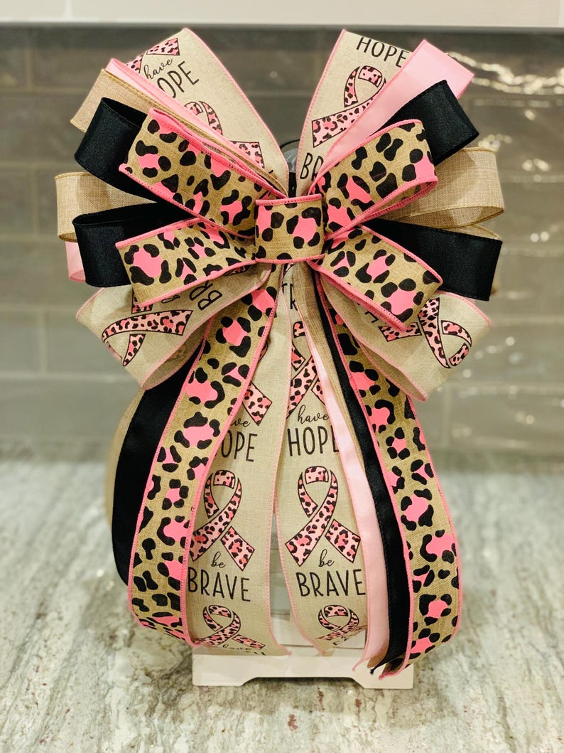 May include: A decorative bow featuring ribbons in pink, black, and tan. The bow has a leopard print pattern in pink and brown. Some ribbons have the words "have HOPE" and "be BRAVE" with a pink ribbon symbol.