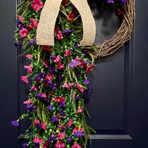 May include: A grapevine wreath adorned with vibrant pink and purple flowers, green foliage, and a burlap bow. The wreath is a decorative piece, perfect for adding a touch of color and natural beauty to any home decor.