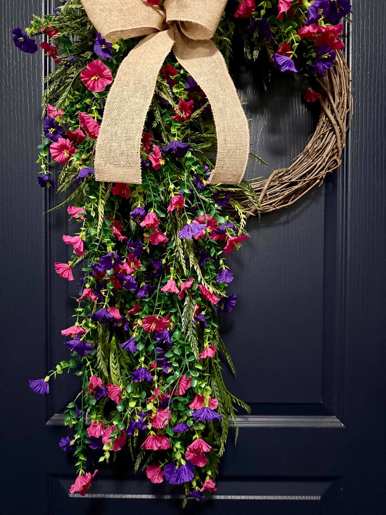 May include: A grapevine wreath adorned with vibrant pink and purple flowers, green foliage, and a burlap bow. The wreath is a decorative piece, perfect for adding a touch of color and natural beauty to any home decor.