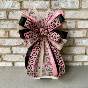 May include: A decorative bow with multiple layers of ribbon in various patterns and colors. The bow features pink, black, and tan ribbons, some with a leopard print design. The bow also includes ribbons with the words "HOPE" and "BE BRAVE".