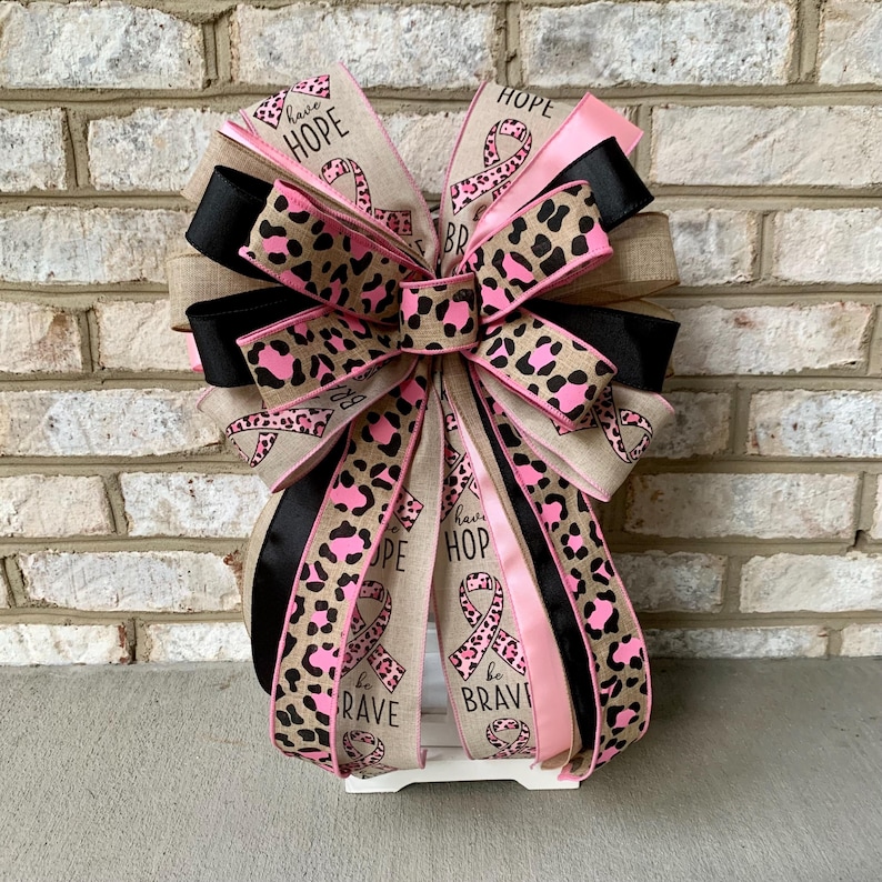 May include: A decorative bow with multiple layers of ribbon in various patterns and colors. The bow features pink, black, and tan ribbons, some with a leopard print design. The bow also includes ribbons with the words "HOPE" and "BE BRAVE".