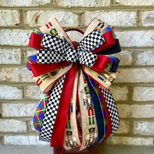 May include: A decorative bow featuring a mix of ribbons in red, black and white checkered, and patterned designs. The bow includes ribbons with nutcracker and plaid patterns. The bow is attached to a red lantern.