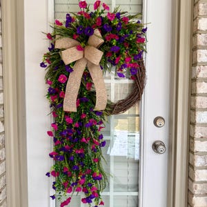 May include: A vibrant floral wreath with a cascading design, featuring pink and purple flowers, green foliage, and a tan burlap bow. The wreath is displayed on a white door with a brick exterior.