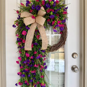 May include: A vibrant floral wreath with a cascading design, featuring a mix of pink and purple flowers, green foliage, and a large burlap bow. The wreath is displayed on a white door, adding a touch of spring decor.