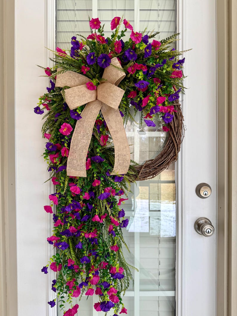 May include: A vibrant floral wreath with a cascading design, featuring a mix of pink and purple flowers, green foliage, and a large burlap bow. The wreath is displayed on a white door, adding a touch of spring decor.