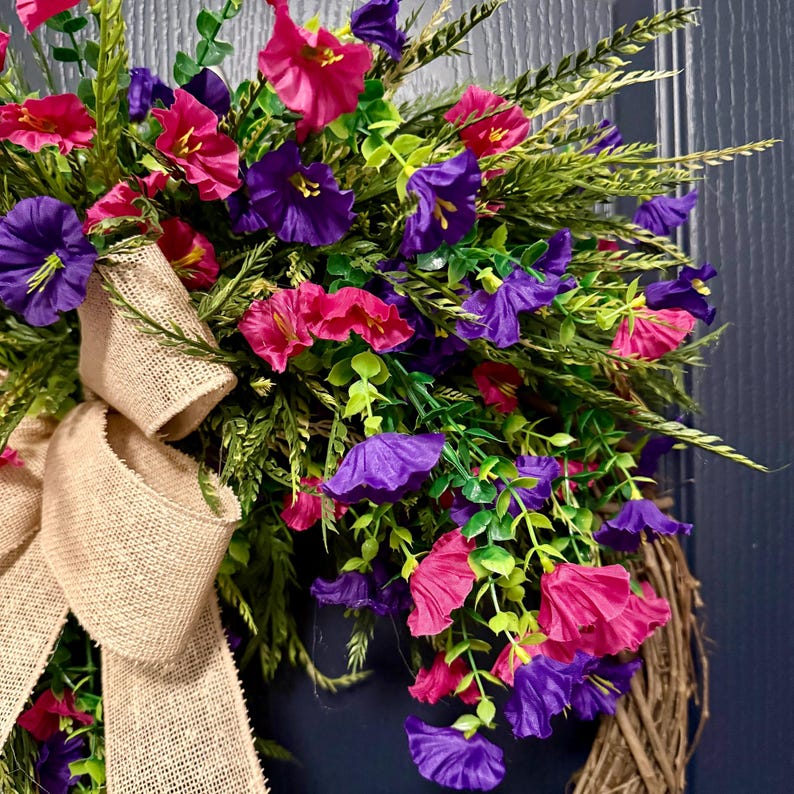 May include: A vibrant floral wreath featuring a mix of pink and purple flowers, green foliage, and wheat-like accents. A large burlap bow adds a rustic touch. The wreath is set against a dark blue background.