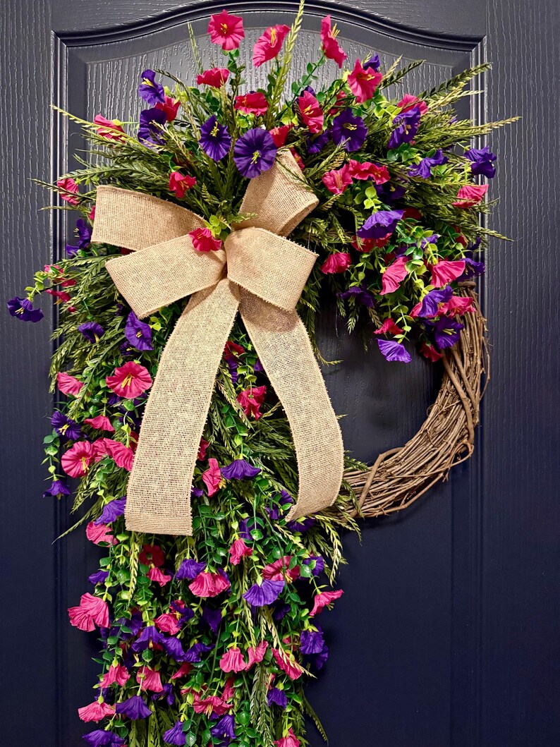 May include: A decorative wreath featuring a natural grapevine base, adorned with vibrant pink and purple flowers, green foliage, and a large burlap bow. This floral wreath is perfect for adding a touch of color to any door or wall.