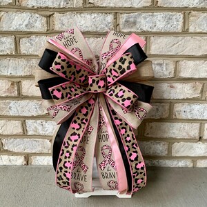 May include: A decorative bow with ribbons in pink, black, and tan. The ribbons feature a leopard print design and the words "HOPE" and "be BRAVE". The bow is likely for a wreath or other decoration.