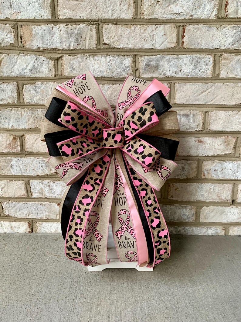 May include: A decorative bow with ribbons in pink, black, and tan. The ribbons feature a leopard print design and the words "HOPE" and "be BRAVE". The bow is likely for a wreath or other decoration.