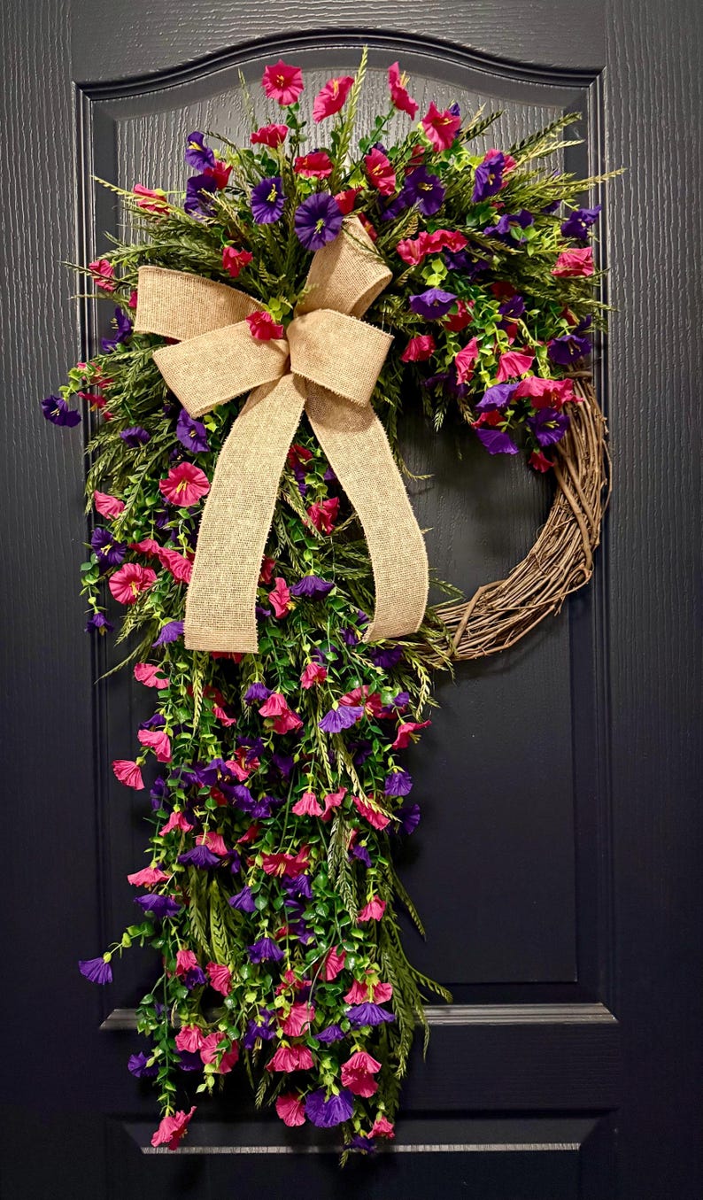 May include: A decorative wreath featuring a natural grapevine base, adorned with vibrant pink and purple flowers, green foliage, and a tan burlap bow. The wreath is designed for door or wall decor.