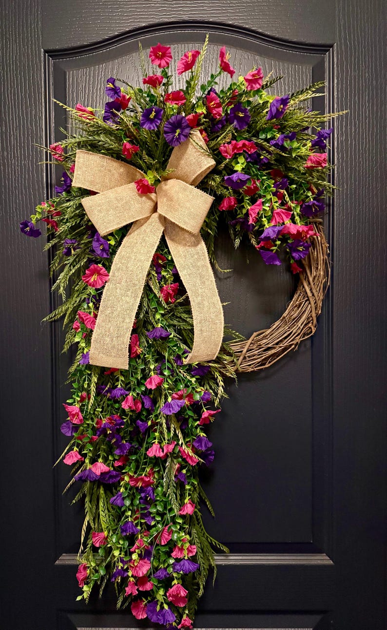 May include: A decorative wreath featuring a natural grapevine base, adorned with vibrant pink and purple flowers, green foliage, and a large burlap bow. The wreath is designed for door or wall decor.